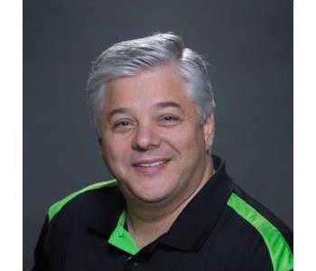 Male employee with gray hair, smiling, wearing a black and green polo shirt, in front of a dark gray background.